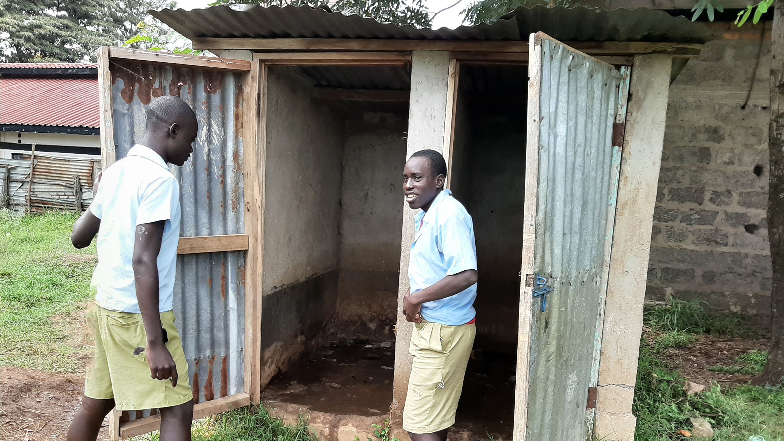 Two boys stand outside a two-door structure made if wood, concrete and iron sheet.