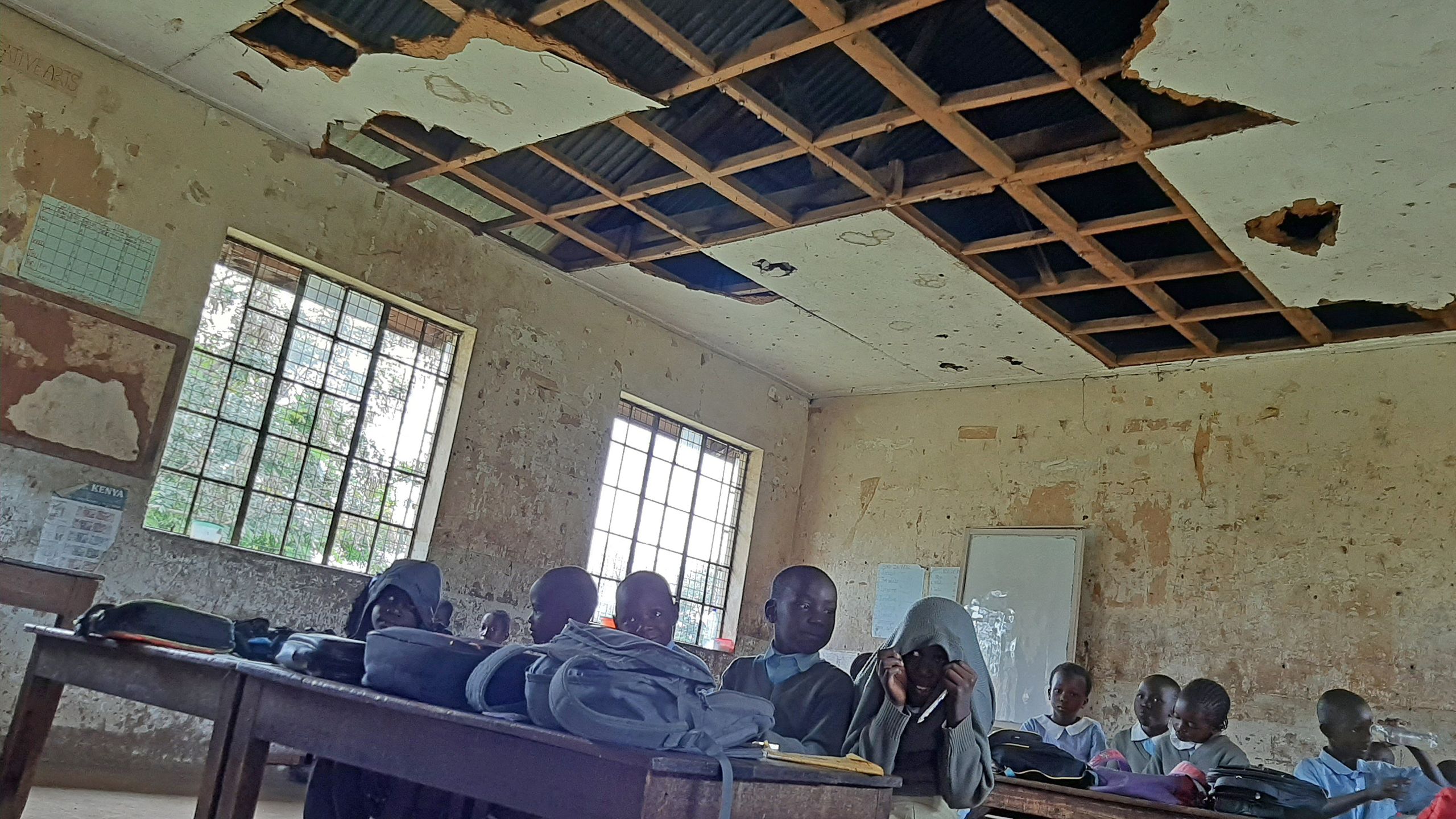 A room with boys and girls working on their desks. The room has a damaged wooden ceiling.