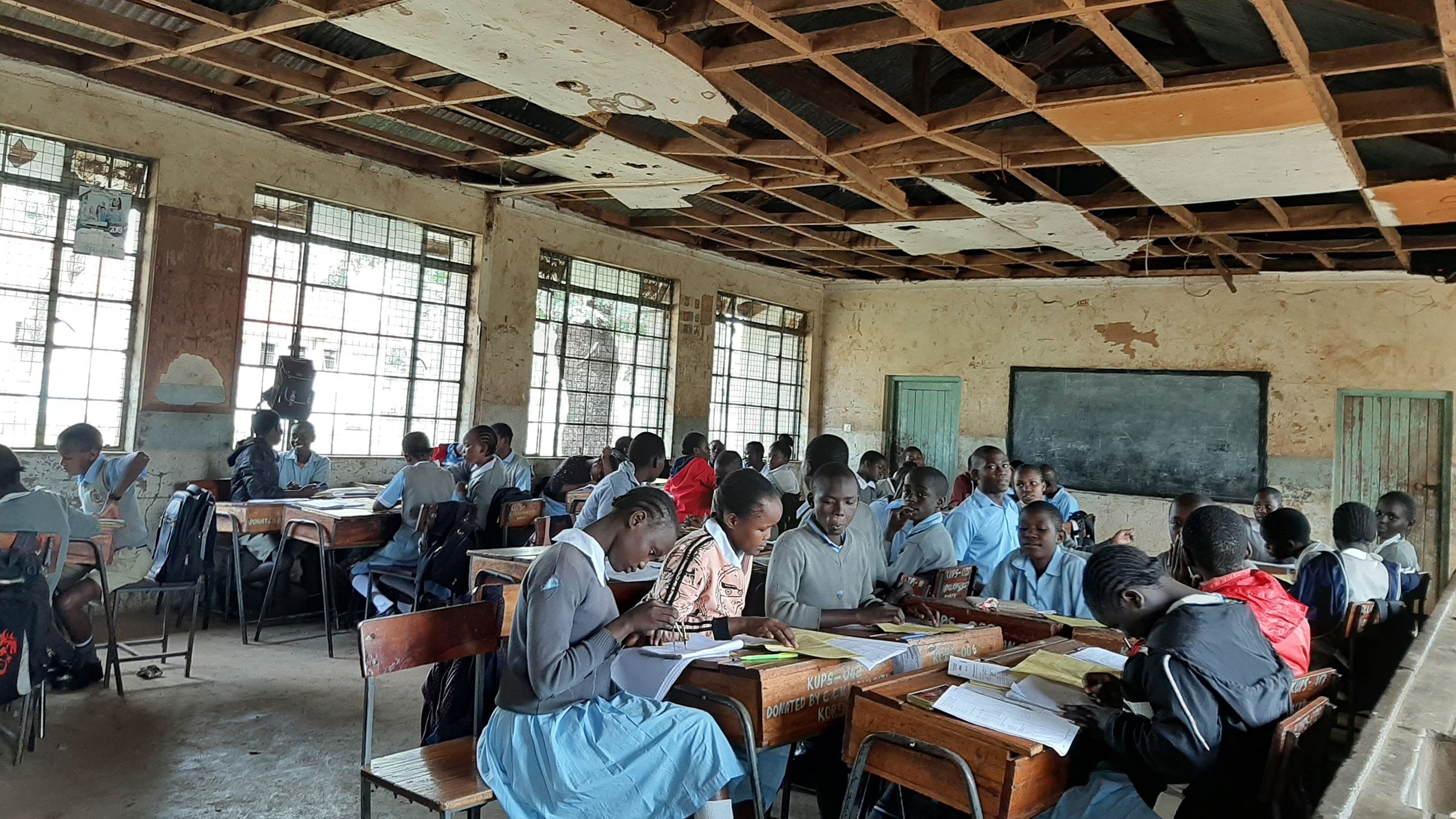 A room with boys and girls working on their desks. The room has a damaged wooden ceiling.